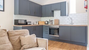 A modern kitchen with grey cabinets, a built-in oven, a microwave, and a toaster. There is a wooden countertop and a beige sofa in the foreground.