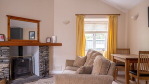 A cozy living room with a stone fireplace, a wooden mantel, a sofa with cushions, a dining table with chairs, and a window with yellow curtains.