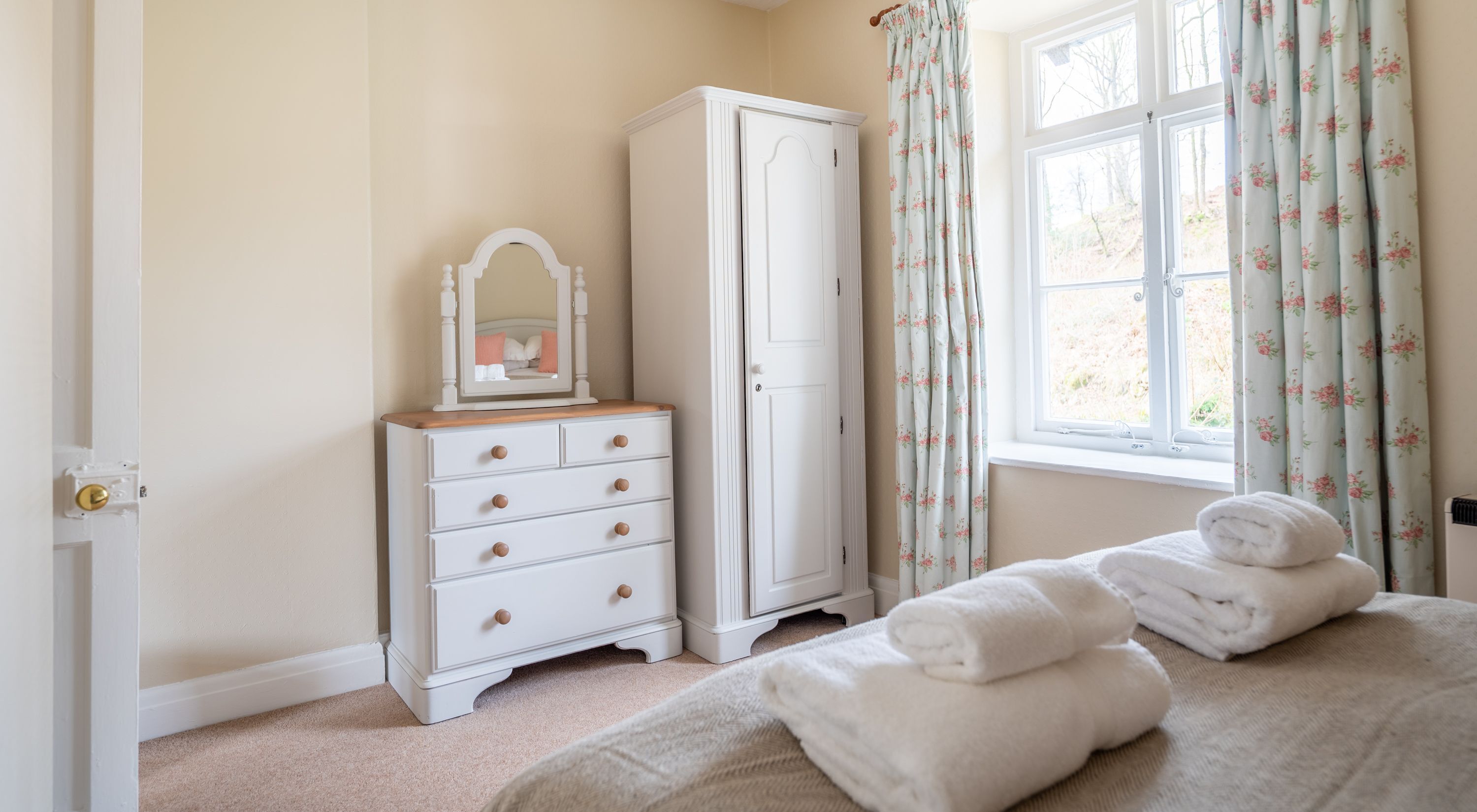 Une chambre avec une commode blanche, une grande armoire, un lit avec des serviettes pliées et une fenêtre ornée de rideaux à motifs floraux.