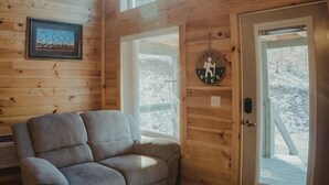 A cozy cabin interior with a gray couch, wooden walls, and large windows offering a view of a snowy landscape. - Secluded 12-Acre Retreat w/ Hot Tub, Mountain Views, Arcade - Red River Gorge (Campton)