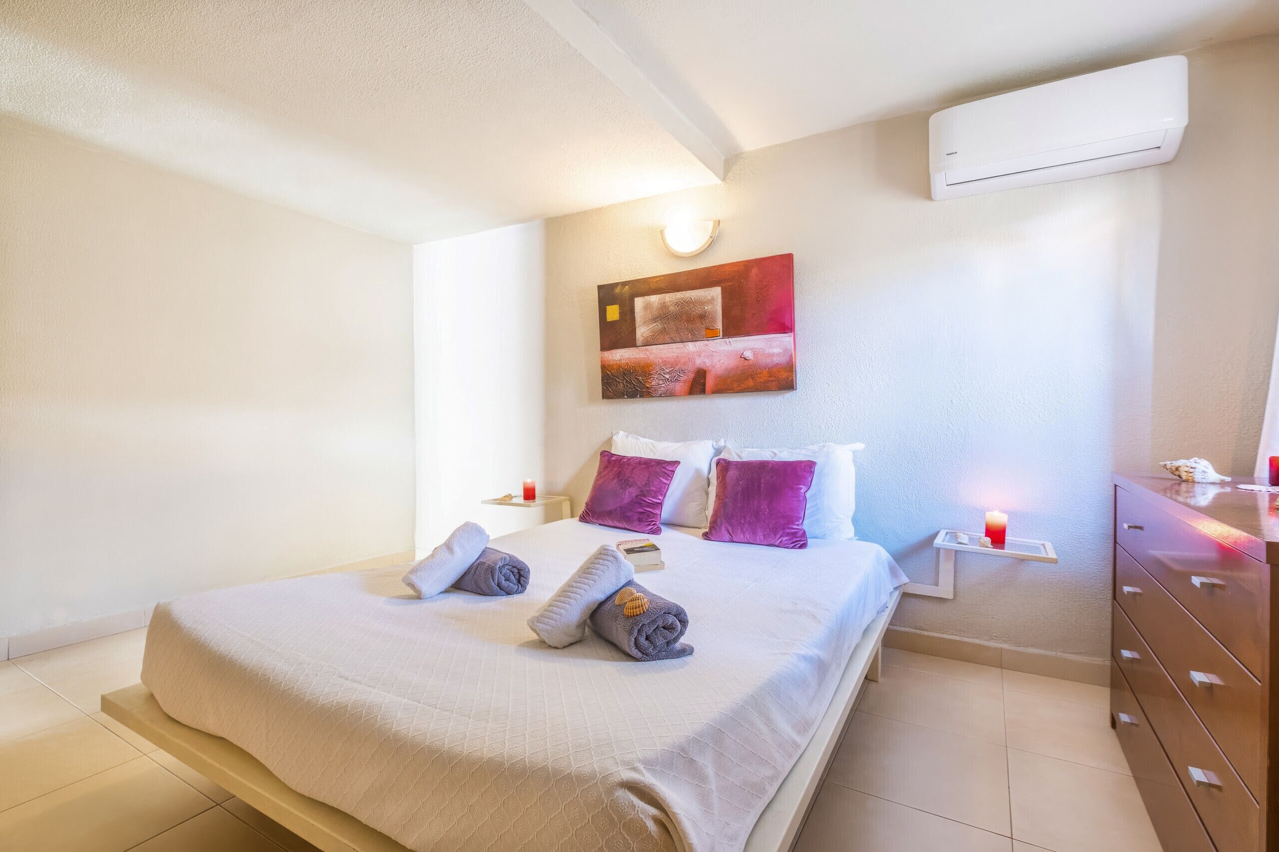 Een hotelkamer met een bed, kussens, een nachtkastje met een kaars, een kunstwerk aan de muur en een airconditioner.