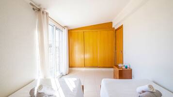 A hotel room with a bed, a wooden wardrobe, a small table with toiletries, and a window with curtains.