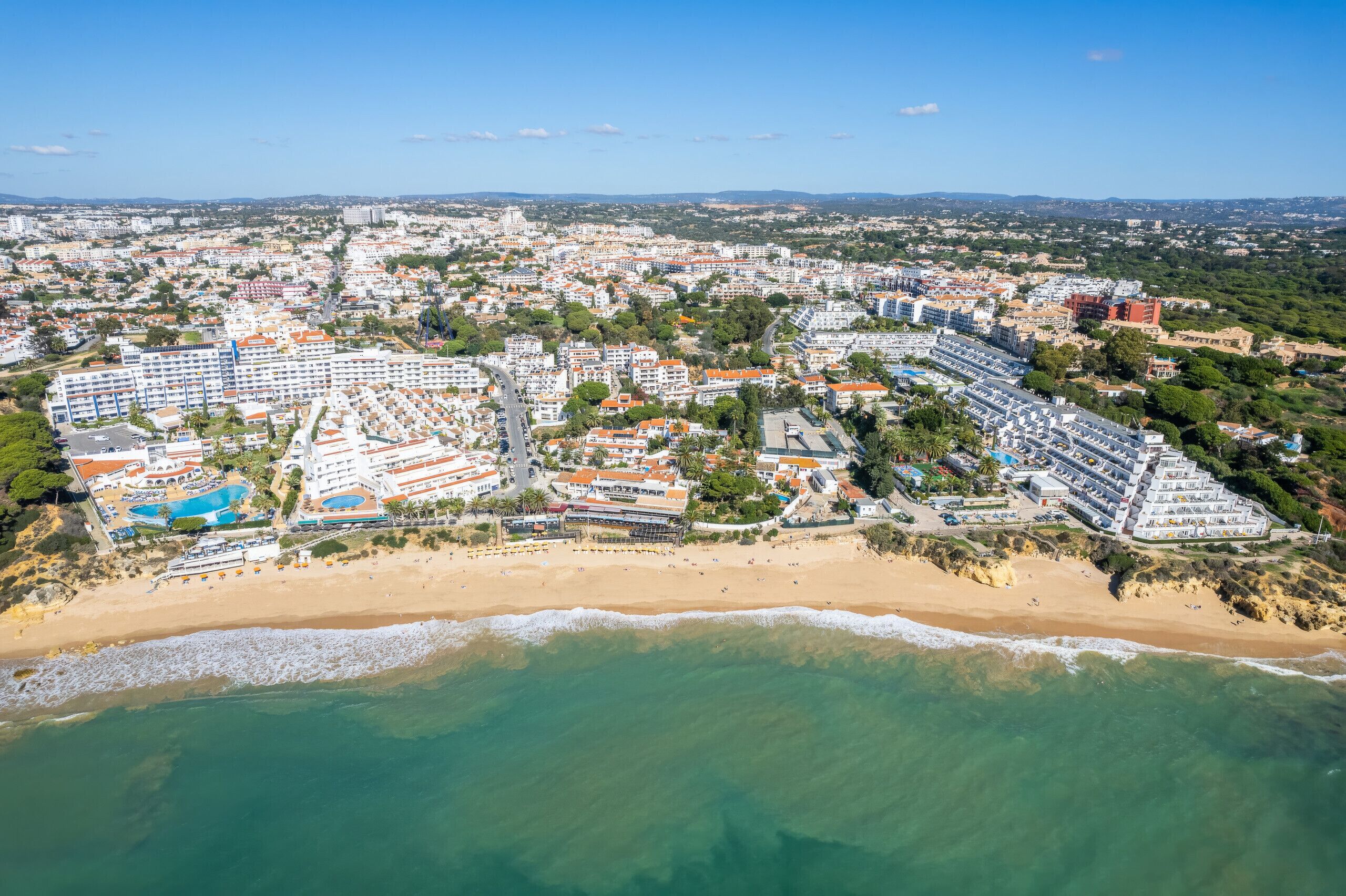 Vista aérea de un complejo turístico costero con varios edificios de hotel, una playa y un cielo azul despejado.