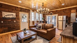 A cozy cabin interior with a wooden ceiling, a leather sofa, a wooden coffee table, and a kitchen area with a stainless steel refrigerator. - Little Blue, cozy cabin with hot tub, fire pit, and cornhole near Polar Express (Bryson City)
