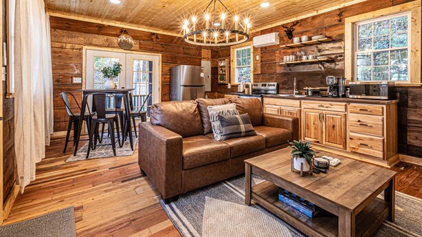 A cozy cabin interior with a wooden ceiling, a chandelier, a leather sofa, a dining area, and a kitchen with wooden cabinets and appliances. - Little Blue, cozy cabin with hot tub, fire pit, and cornhole near Polar Express (Bryson City)