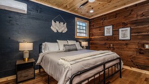 A bedroom with a bed, bedside lamps, a ceiling fan, a window, and wooden walls. - Little Blue, cozy cabin with hot tub, fire pit, and cornhole near Polar Express (Bryson City)