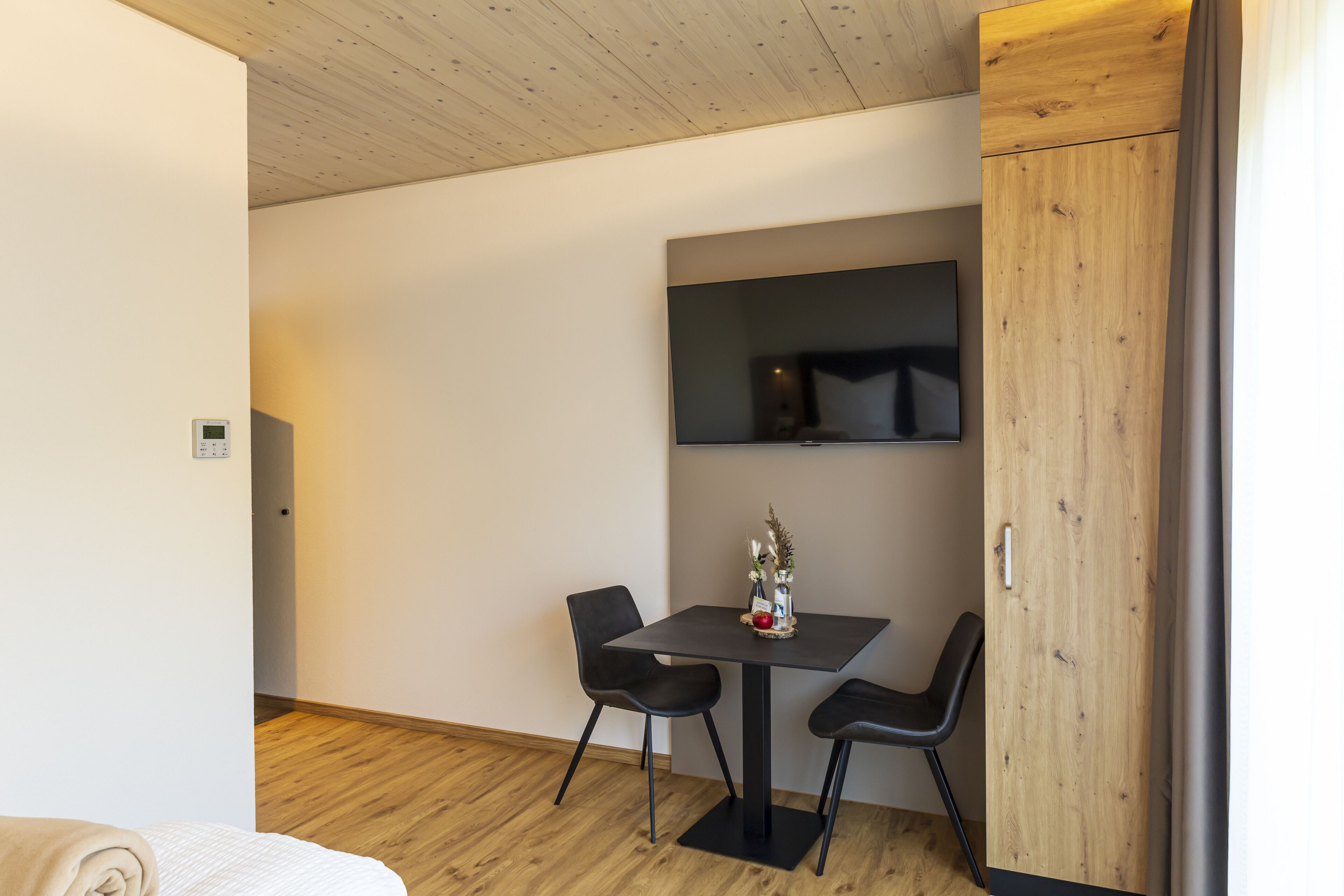 A small, modern room with a wooden ceiling, a flat-screen TV mounted on the wall, a small table with two chairs, and a wooden wardrobe.