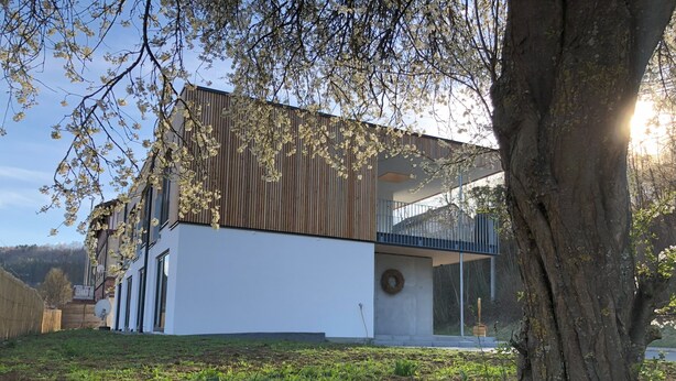 Una casa moderna de dos pisos con balcón, rodeada de follaje y un árbol grande.