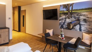 Una habitación moderna con suelo de madera, una cama, una mesa de comedor con sillas, un televisor montado en la pared y un cuadro de un paisaje.