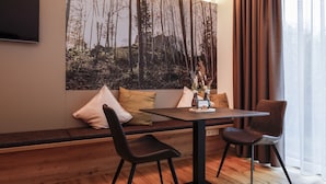 Una habitación moderna con suelo de madera, un sofá con cojines, una mesa de comedor con sillas y un cuadro grande con motivos forestales en la pared.