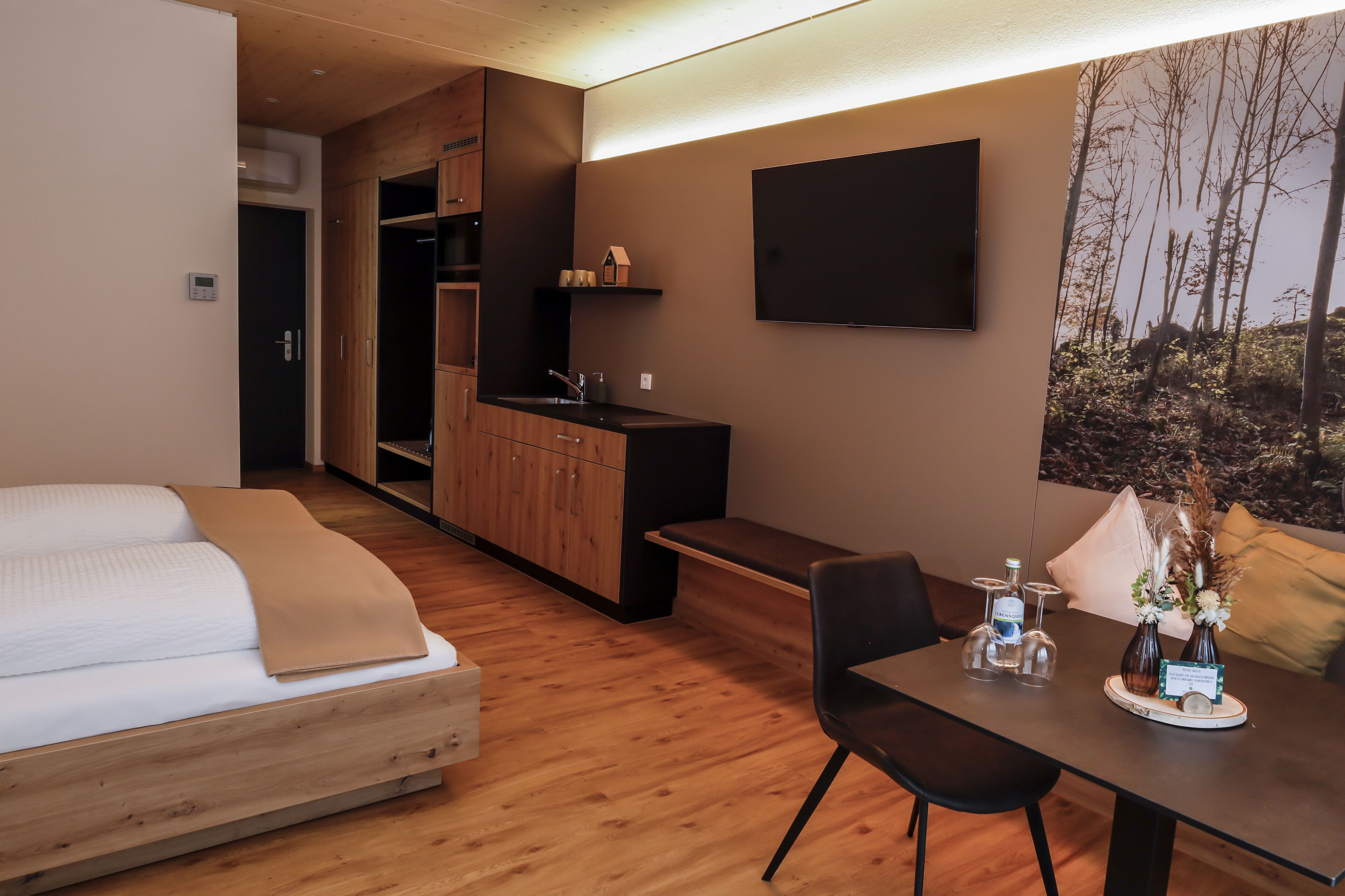 A modern hotel room with a bed, a dining table with chairs, a kitchen area, and a flat-screen TV mounted on the wall.
