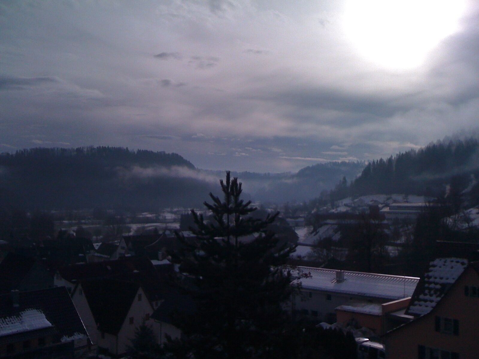 A residential area with houses, some with snow on the roofs, and a forested hill in the background.