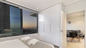 Une chambre moderne dotée d’un grand lit, offrant une vue sur la ville grâce à de grandes fenêtres du sol au plafond, et comprenant un coin repas séparé avec une table et des chaises.