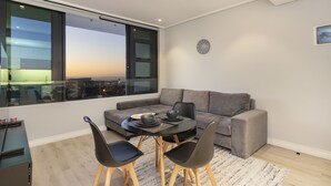 Un salon moderne avec un canapé gris, une table à manger et des chaises, et une vue sur la ville grâce à de grandes fenêtres.