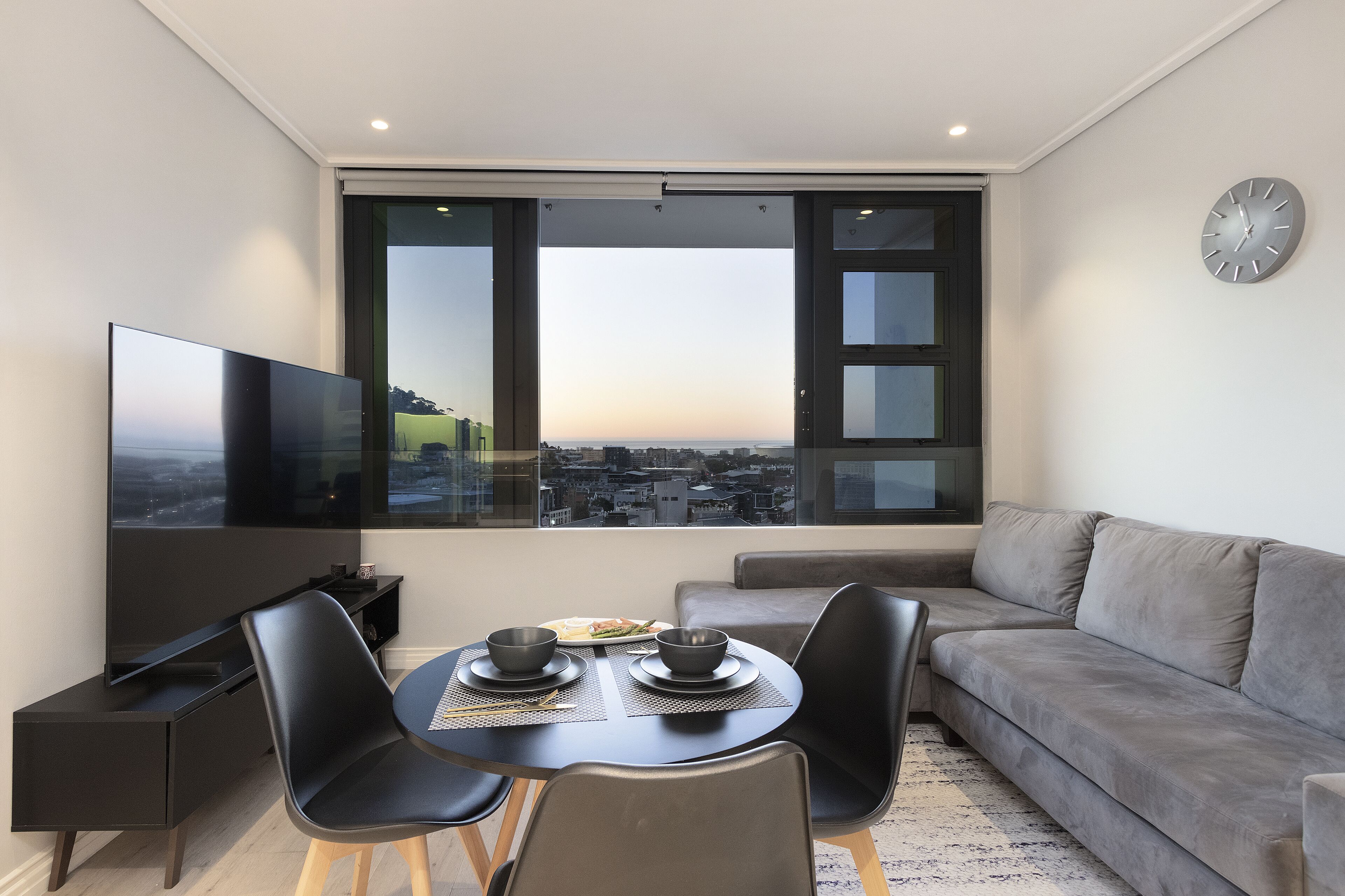 Un salon moderne avec un téléviseur à écran plat, une table à manger avec deux chaises et un canapé gris.