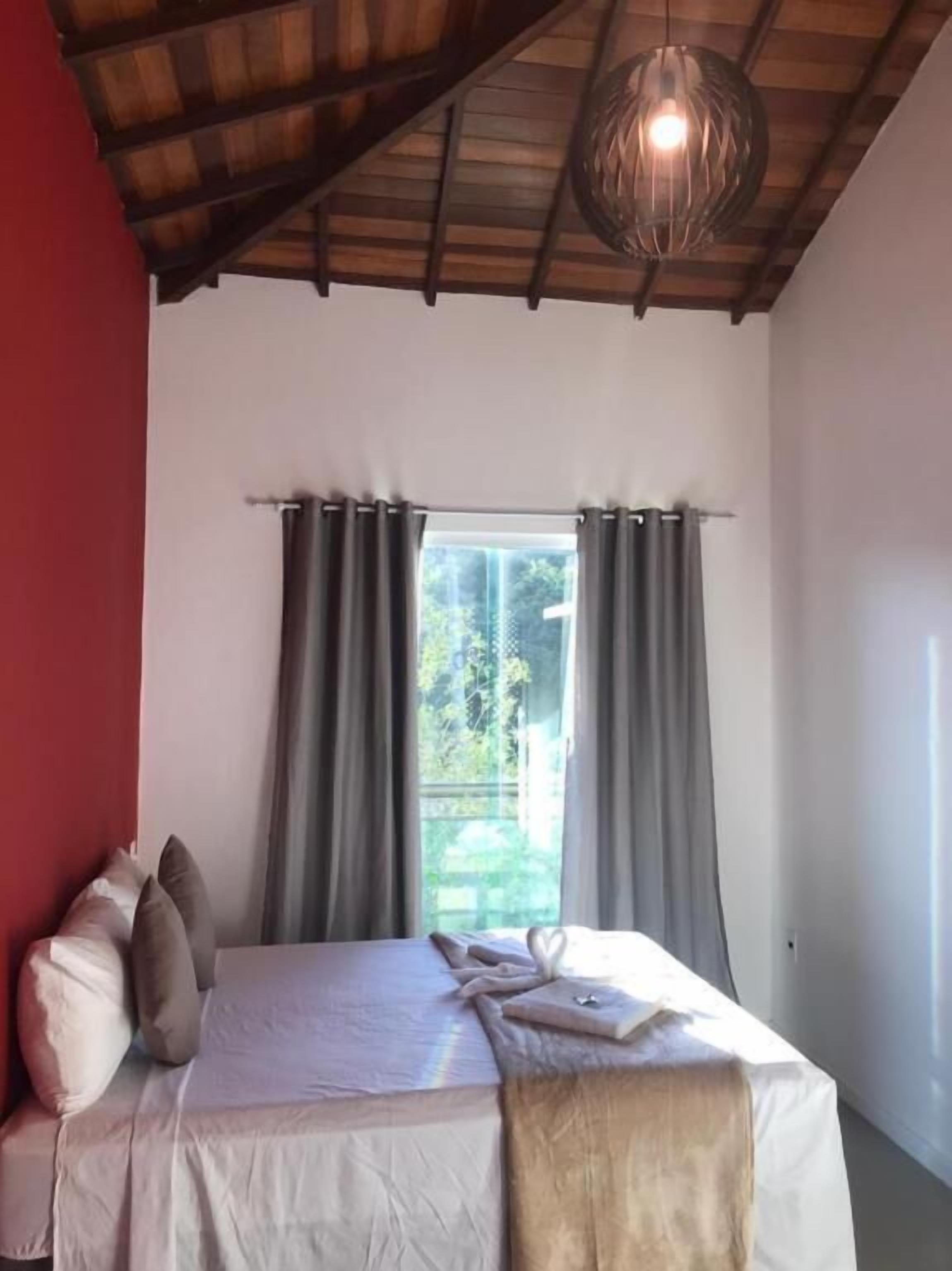 A bedroom with a bed, a window with curtains, a wooden ceiling, and a hanging light fixture.