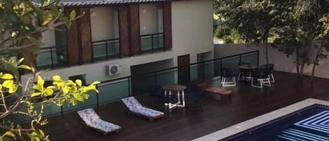 A pool area with lounge chairs and a dining table, surrounded by greenery and a hillside in the background.