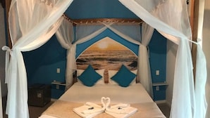 A bed with a canopy, a painting of a beach on the headboard, and two towels arranged in a heart shape on the bed. - Foca Guest House (Búzios)