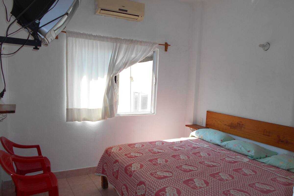 Un dormitorio con cama, ventana con cortinas, una silla roja y aire acondicionado instalado en la pared.