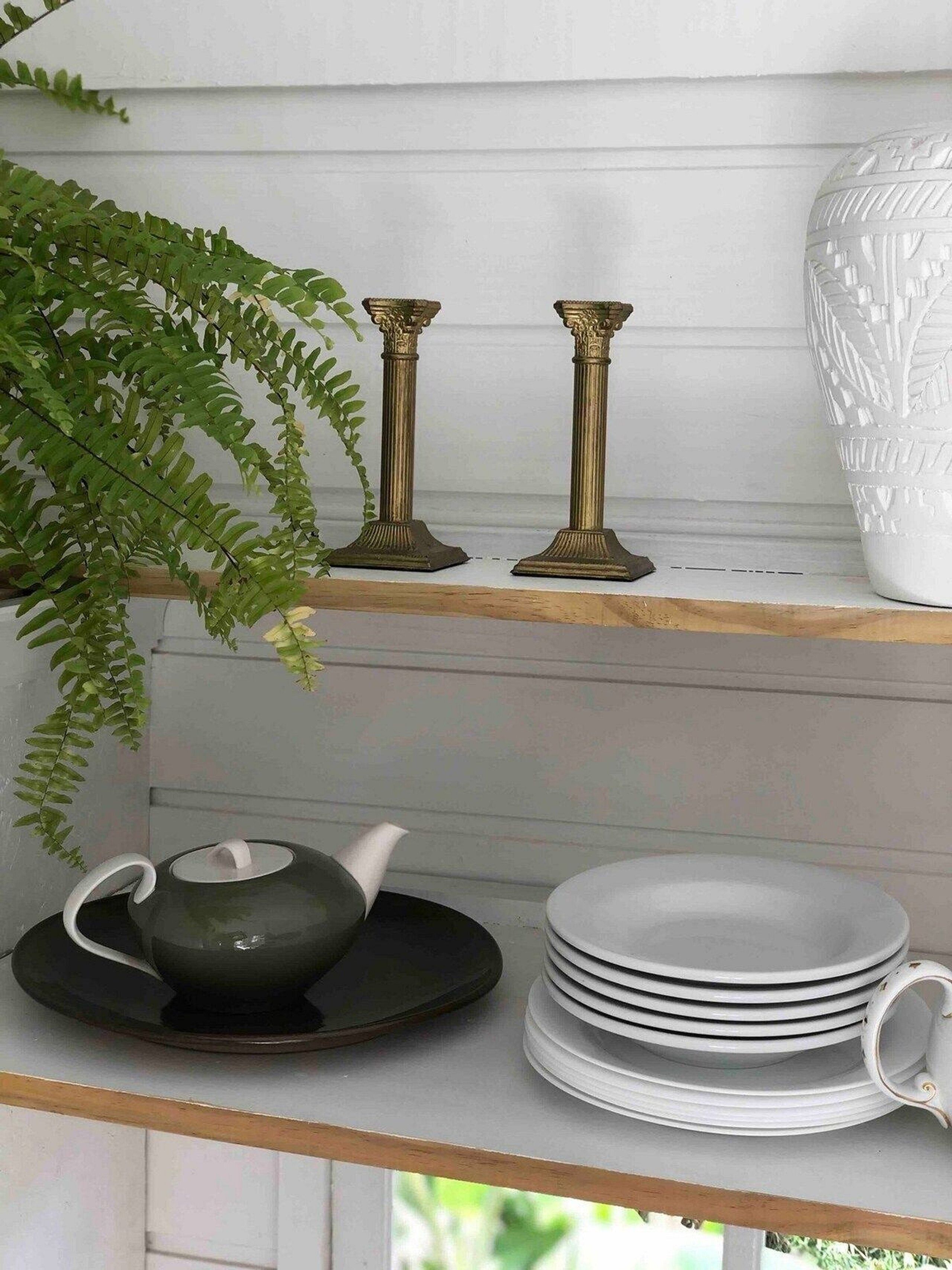 A shelf with two brass candle holders, a white vase, a green teapot, and stacked white plates.