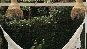 A hammock suspended between two poles, with a metal roof overhead and a lush green hedge in the background. - Chalé Super Charmoso - CasaIvan (Florianópolis)