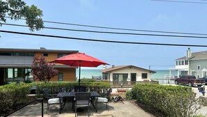 Une terrasse avec des meubles d’extérieur et un parasol rouge, d’où l’on peut admirer une plage et une maison à vendre.