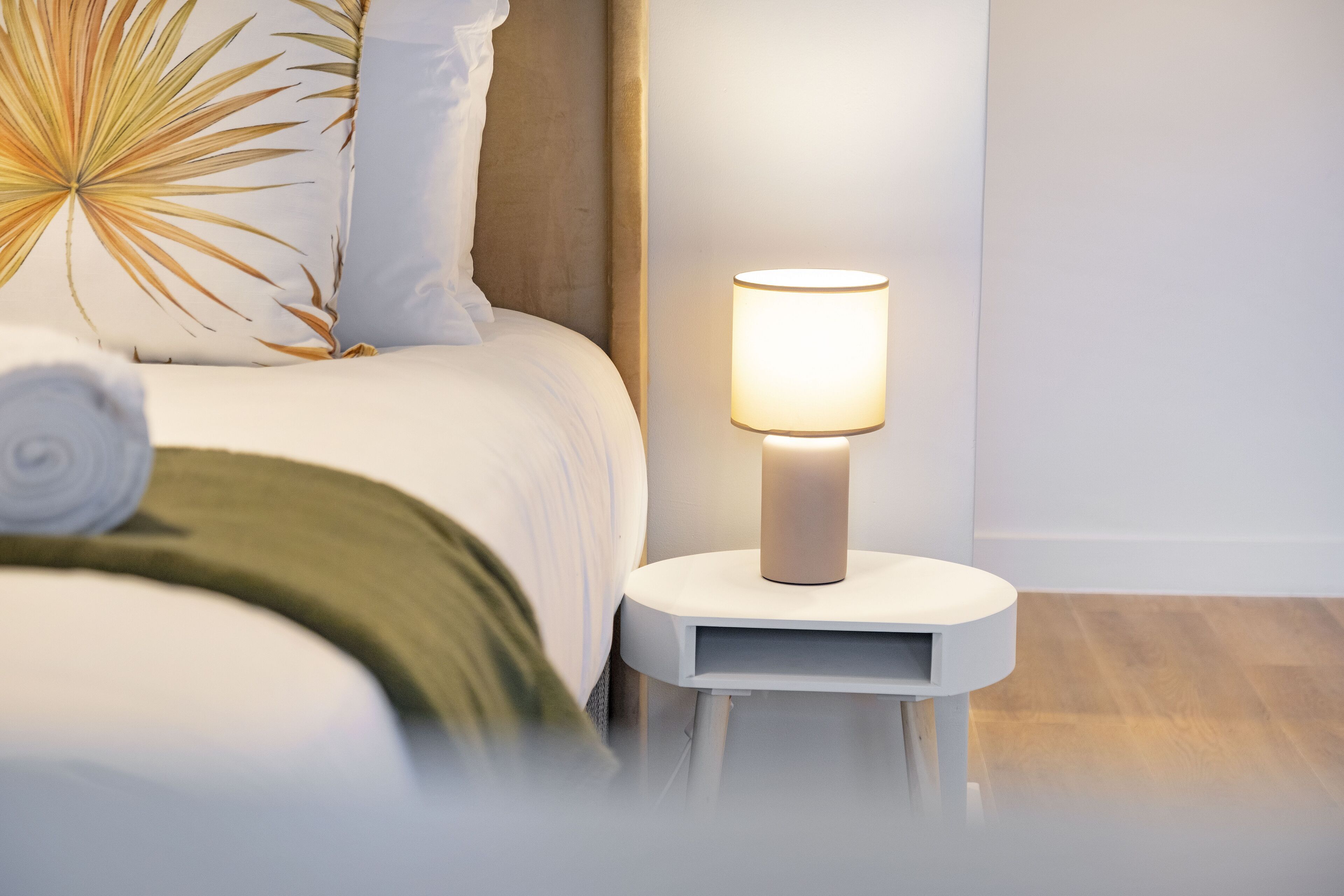 Ein ordentlich bezogenes Bett mit einem zusammengerollten Handtuch, einer Nachttischlampe und einem Kissen aus Palmblatt.
