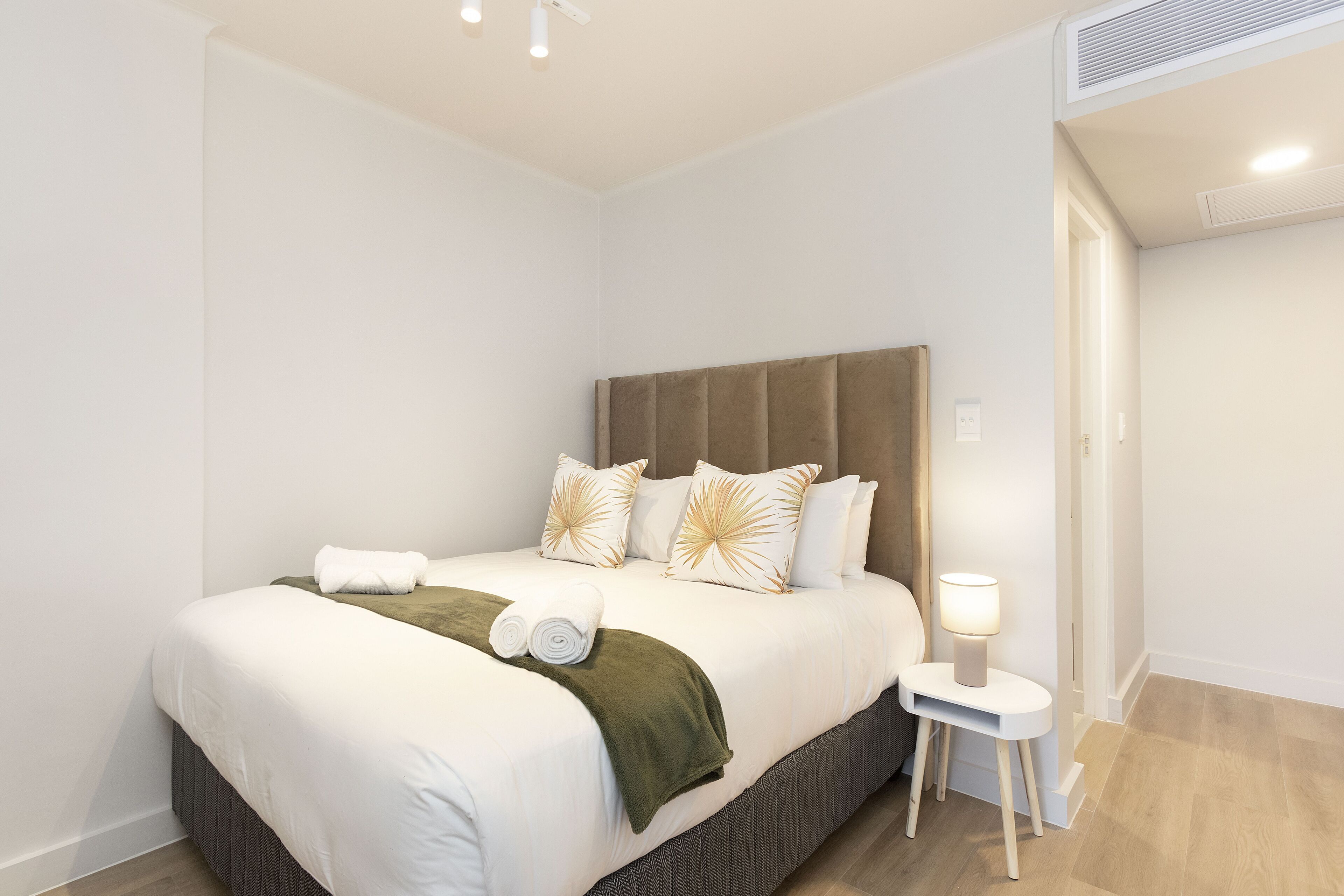 Ein ordentlich bezogenes Bett mit weißen Leinentüchern, einer grünen Tagesdecke und dekorativen Kissen. Ein kleiner Nachttisch mit einer Lampe ist sichtbar.