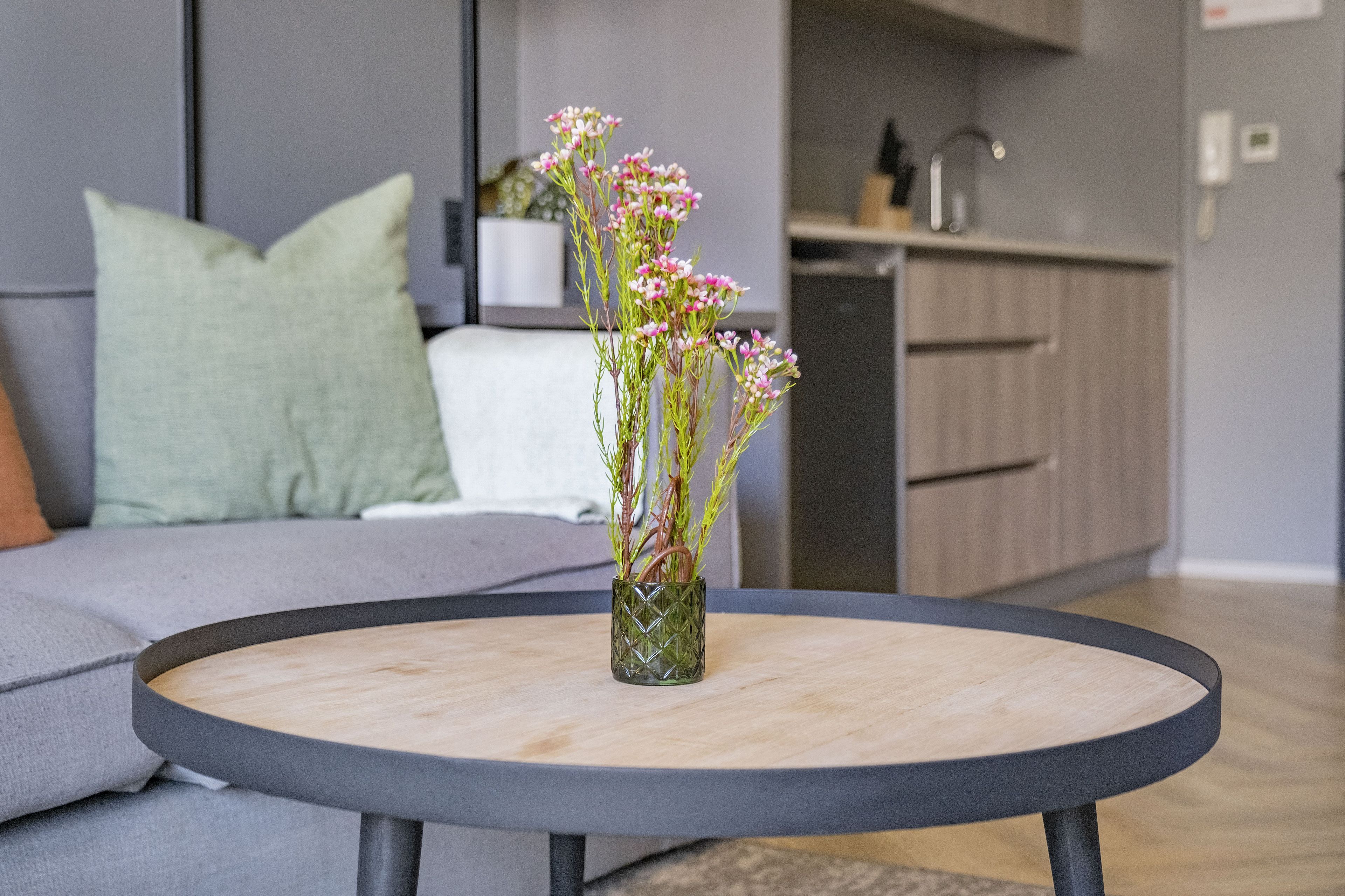 Una mesa redonda con un jarrón de flores en el centro, un sofá gris con un cojín verde y una zona de cocina al fondo.