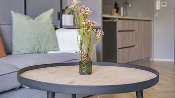 Una mesa redonda con un jarrón de flores en el centro, un sofá gris con un cojín verde y una zona de cocina al fondo.