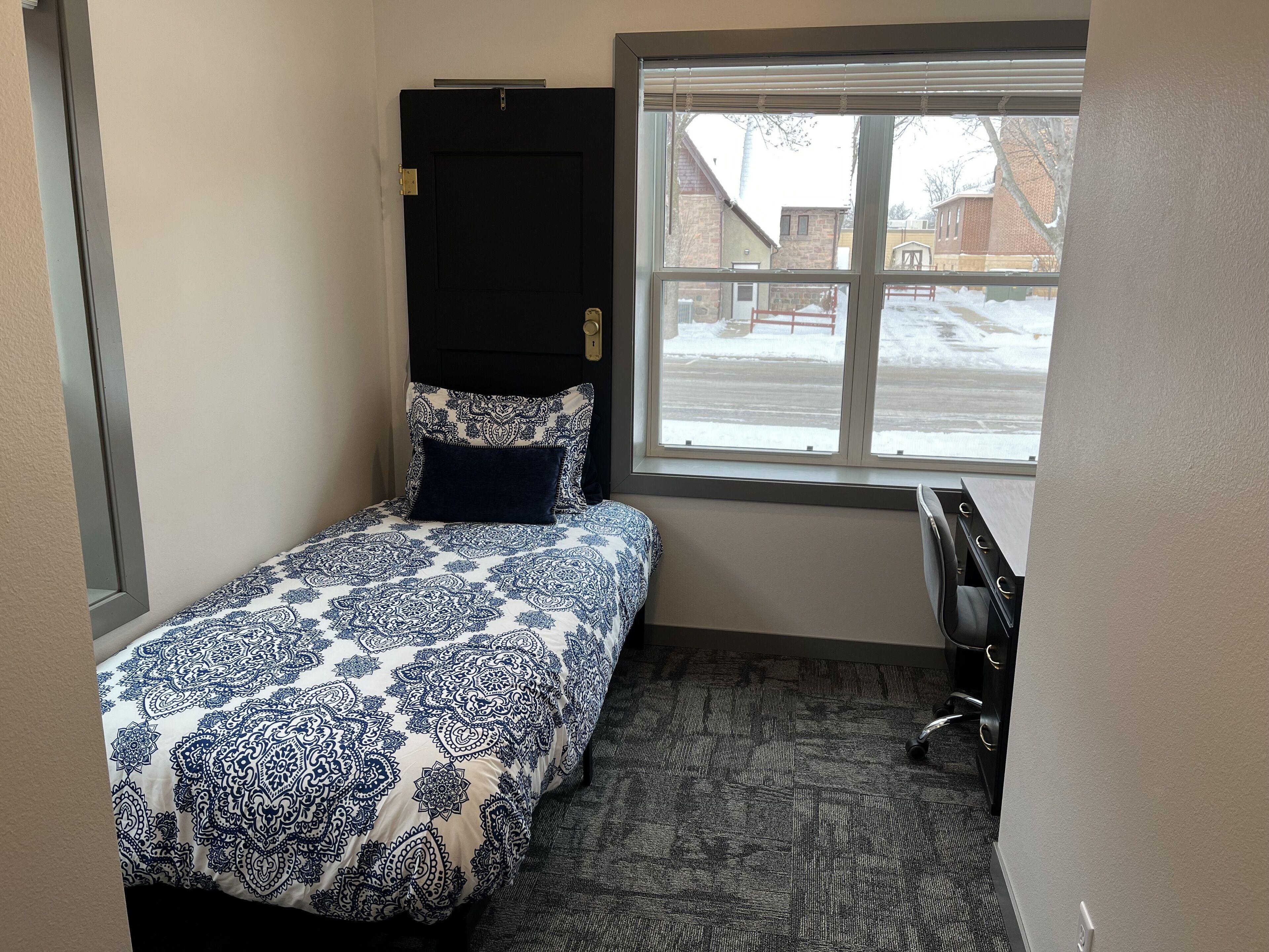 A small, compact bedroom with a single bed, a desk, and a window providing a view of the outside.