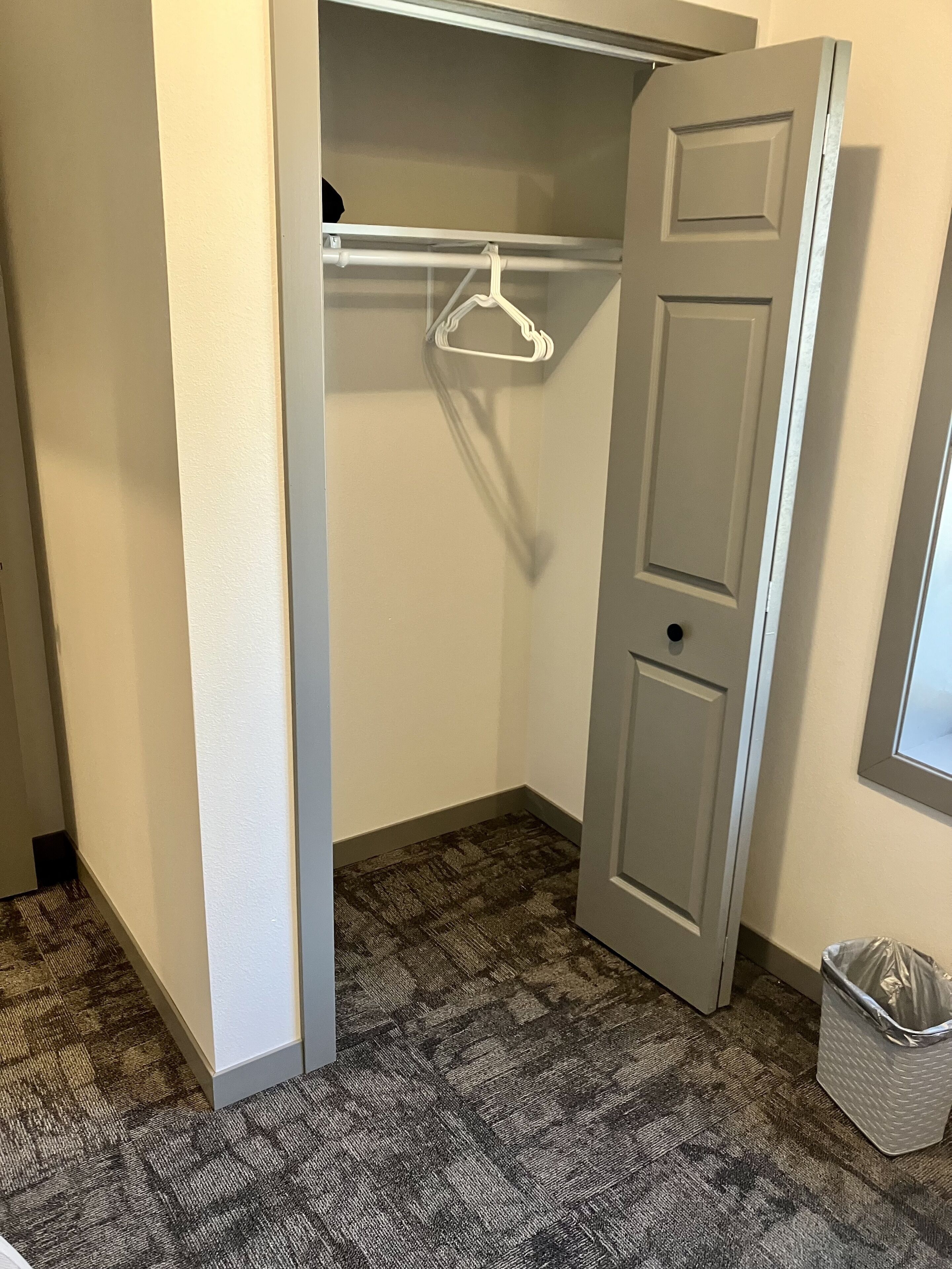 Una habitación pequeña y estrecha, con alfombra gris, un armario con una percha blanca y una puerta gris.