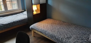 Quarto tipo dormitório com duas camas individuais, uma mesa de cabeceira com luminária, uma cadeira e uma janela.