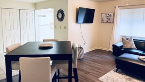 A living room with a dark wooden dining table, beige chairs, a black leather sofa, and a television mounted on the wall. - Beautiful 2 bedroom unit across from UF stadium
 (Gainesville)