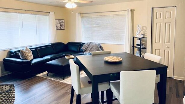 A living room with a black sectional sofa, a dark wooden dining table with white chairs, and a ceiling fan. - Beautiful 2 bedroom unit across from UF stadium
 (Gainesville)