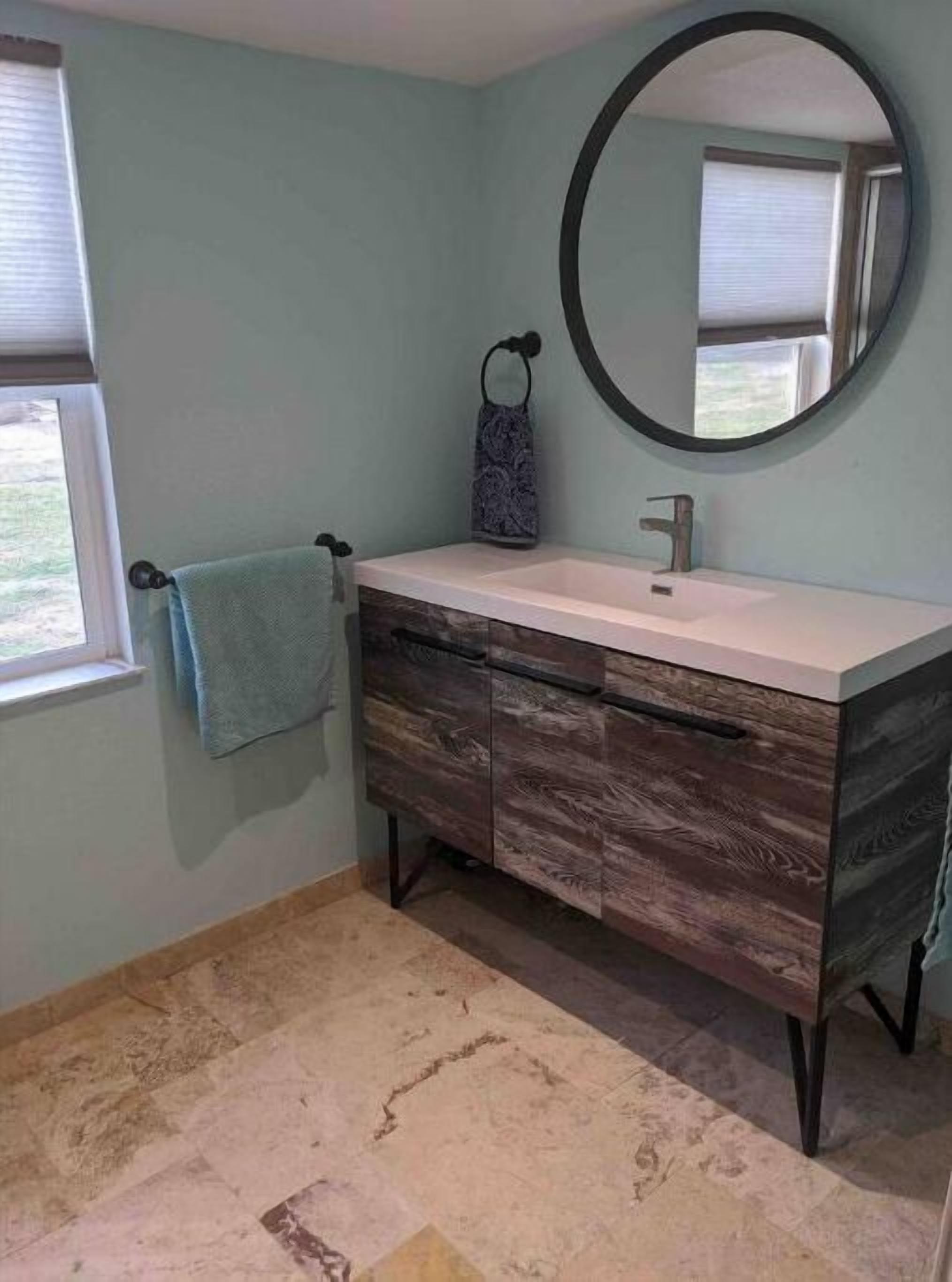 A bathroom with a vanity, a round mirror, a towel rack, and a window.