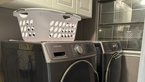 Two front-loading washing machines stacked on top of each other in a laundry room with cabinets above and a window to the right.