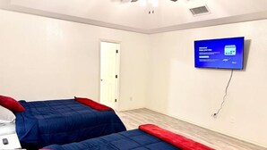 A hotel room with two beds, each with a blue bedspread and red pillows. There is a flat-screen TV mounted on the wall, and a ceiling fan above the beds.