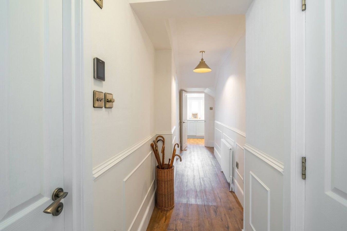 A narrow hallway with white walls, a wooden floor, a basket with walking sticks, and a door with a brass handle.
