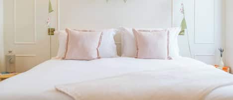 Un letto rifatto con biancheria e cuscini bianchi, un comodino con una lampada e una ghirlanda floreale appesa al muro.