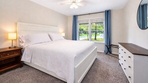 Ein Schlafzimmer mit Bett, Nachttisch, Schminktisch, Deckenventilator und einem Fenster mit Vorhängen.