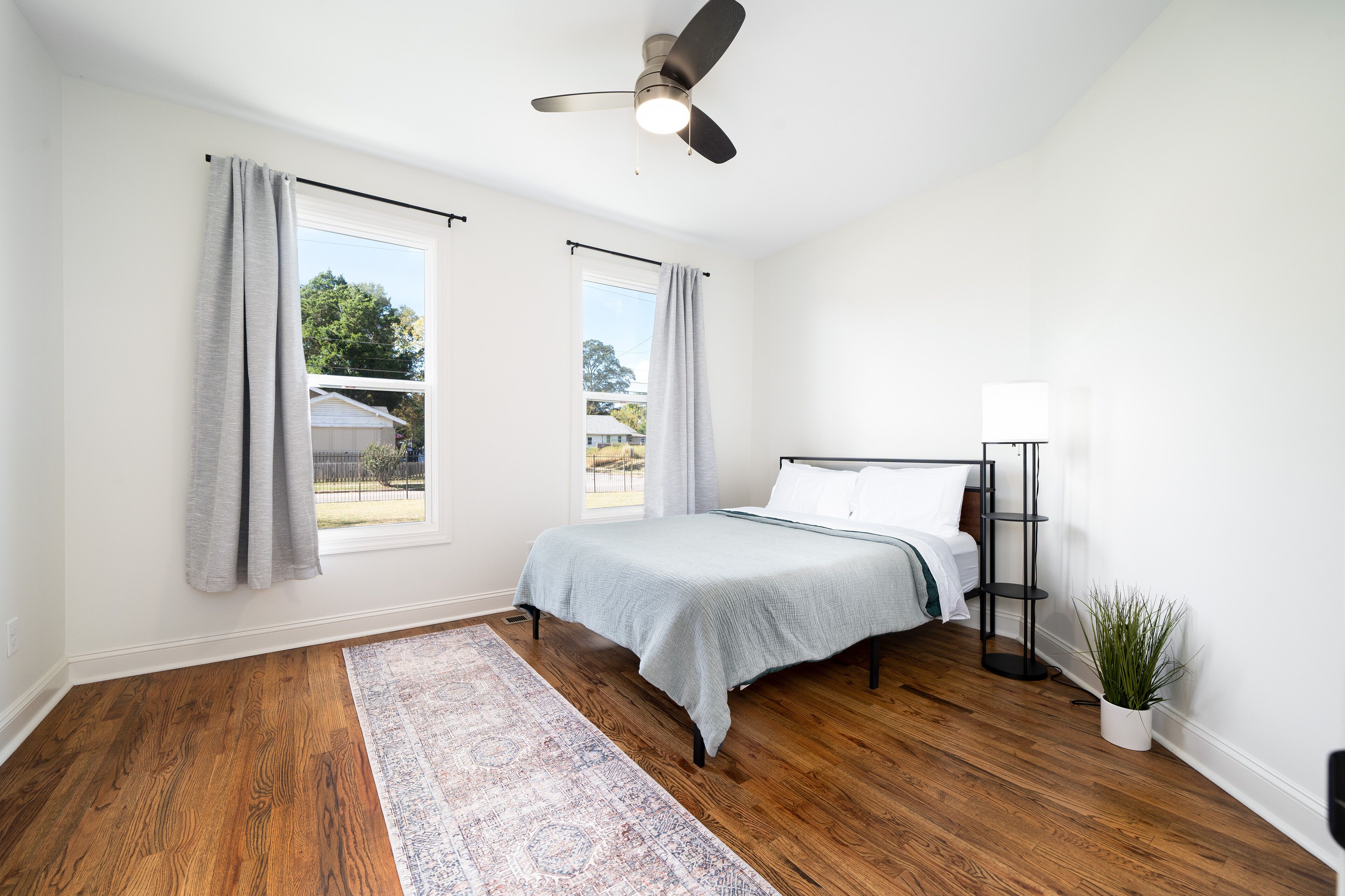 Un dormitorio con cama, lámpara de pie, ventilador de techo y ventana con cortinas.