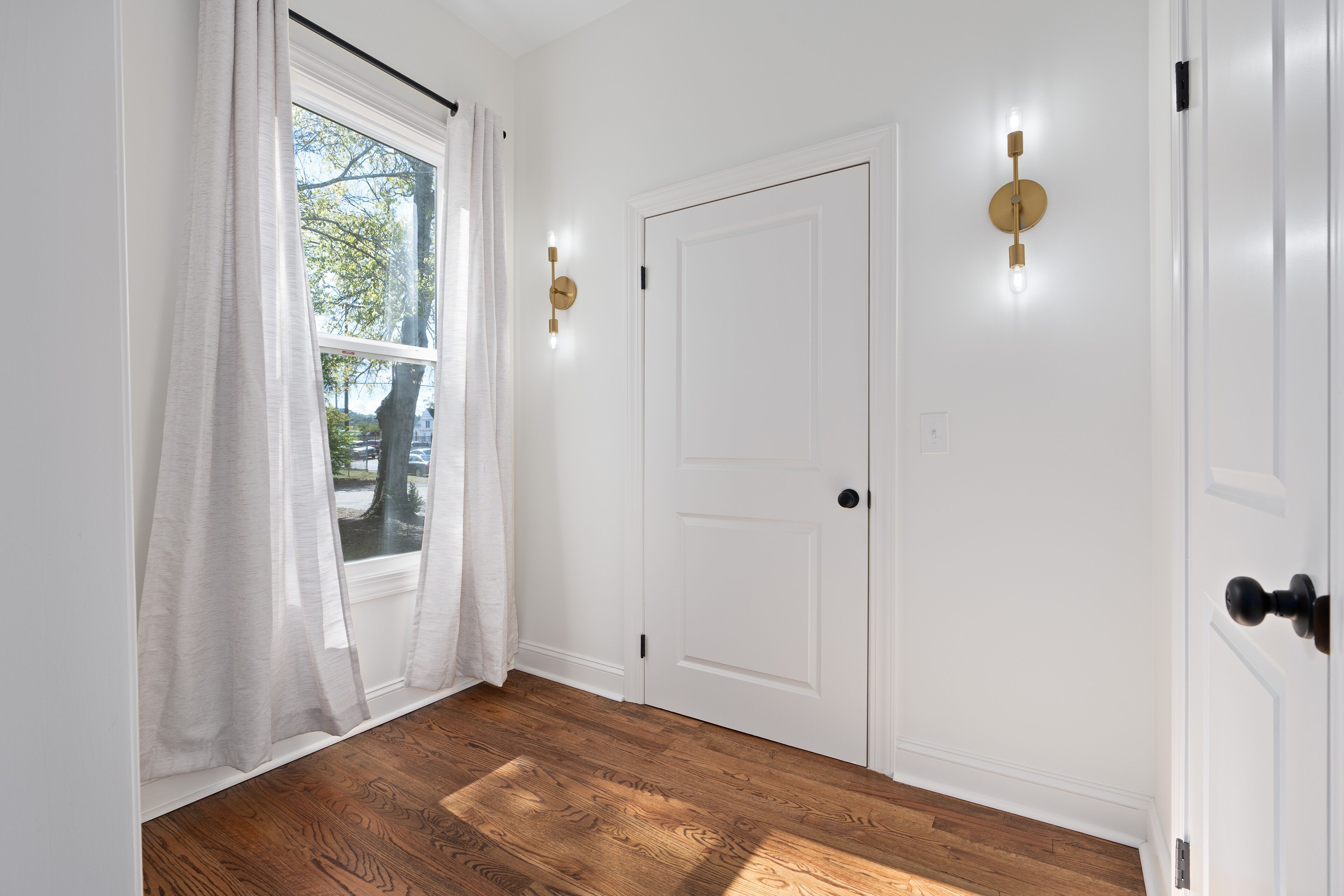 Un pasillo con una puerta blanca, una ventana con cortinas translúcidas y una lámpara fijada a la pared.