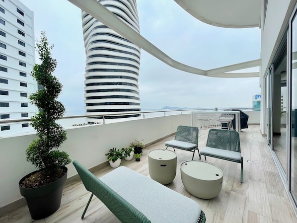 Uma varanda moderna com vista para um edifício alto, plantas em vasos e área de estar externa.