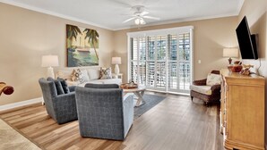 Ein Wohnzimmer mit Deckenventilator, einem Strandbild, einem Sofa, einem Couchtisch und einem an der Wand montierten Fernseher.