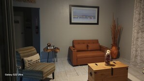 A living room with a brown sofa, a small table with a clock, a chair with a cushion, a vase with decorative sticks, and a framed picture on the wall. - La Posita Beach House  (Cabo Rojo)