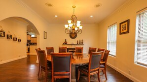 Una sala da pranzo con un tavolo rotondo in legno circondato da sei sedie, un lampadario e una cucina visibile attraverso un arco.