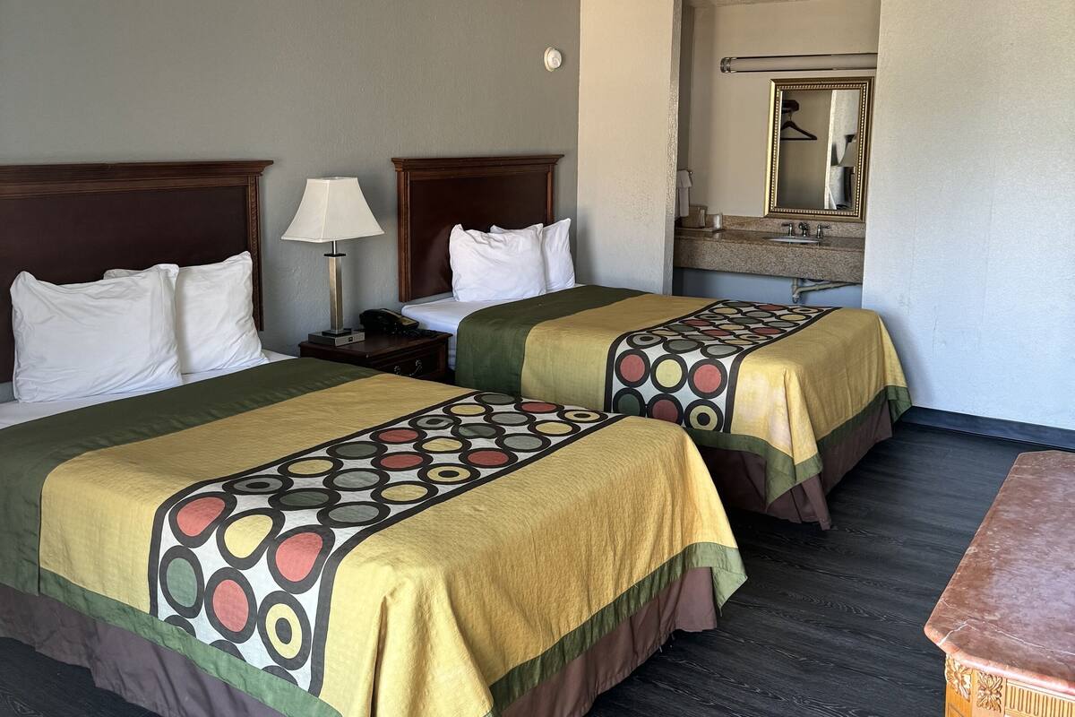 A hotel room with two beds, a nightstand, a table, a mirror, and a bathroom visible in the background.