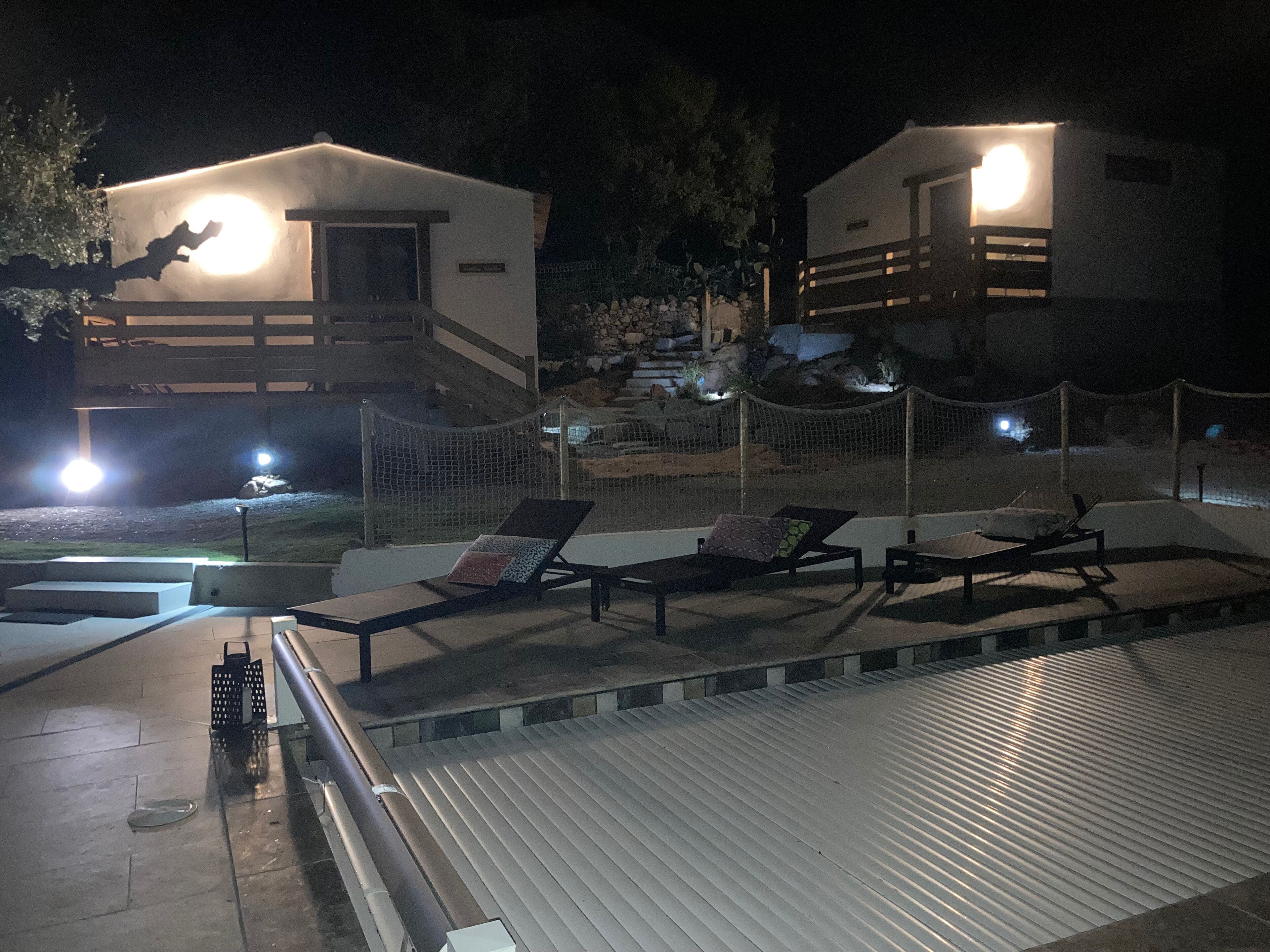 A nighttime view of a resort with two guest houses, a pool area with lounge chairs, and a stone wall.