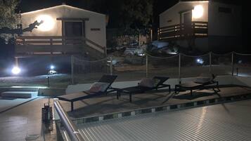 A nighttime view of a resort with two guest houses, a pool area with lounge chairs, and a stone wall.